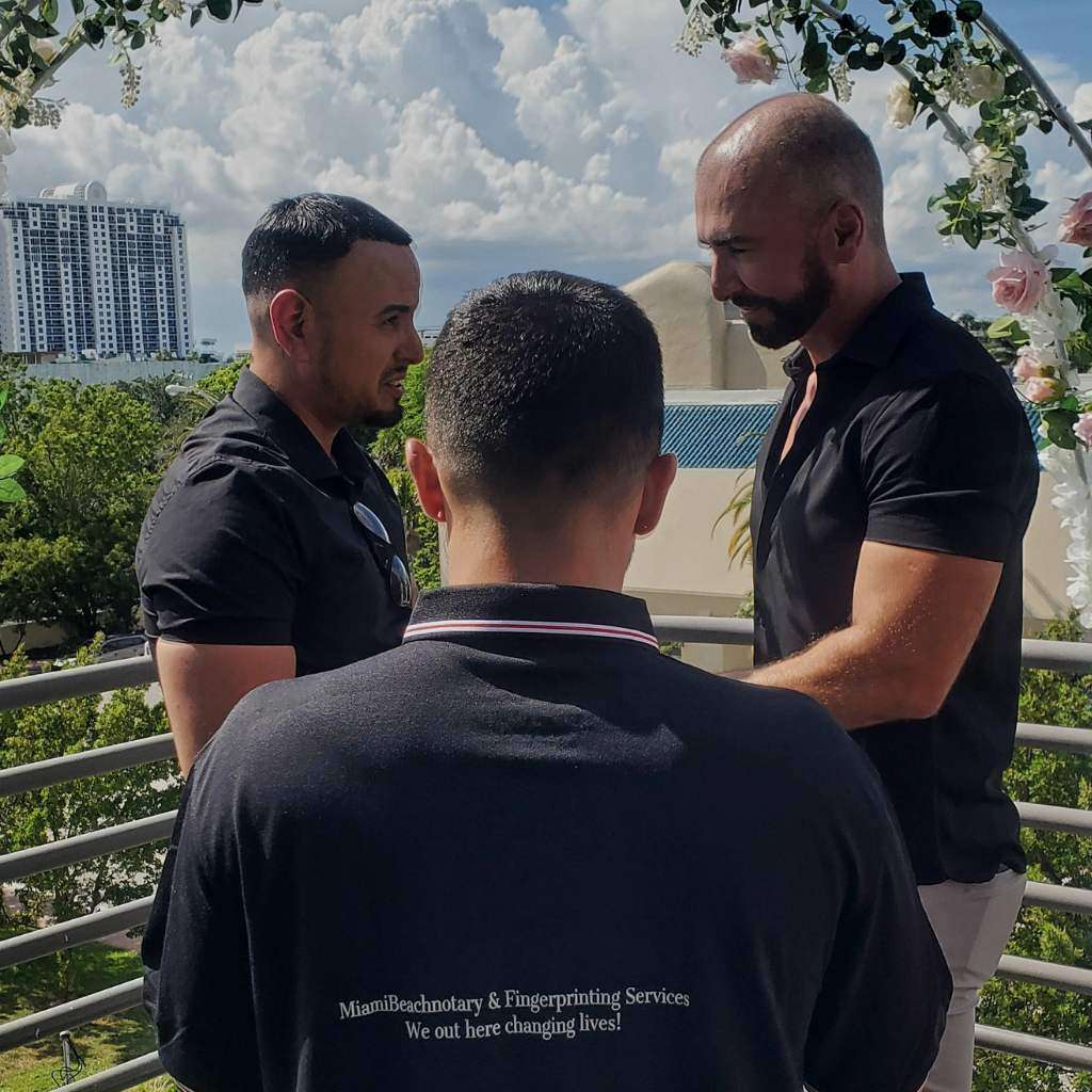 Three men standing on an outdoor balcony with a cityscape and foliage in the background. Two of the men are facing each other, holding hands, celebrating their same-sex marriage, while the third man, seen from behind, appears to be officiating. The officiant's shirt reads "MiamiBeachnotary & Marriage Services.