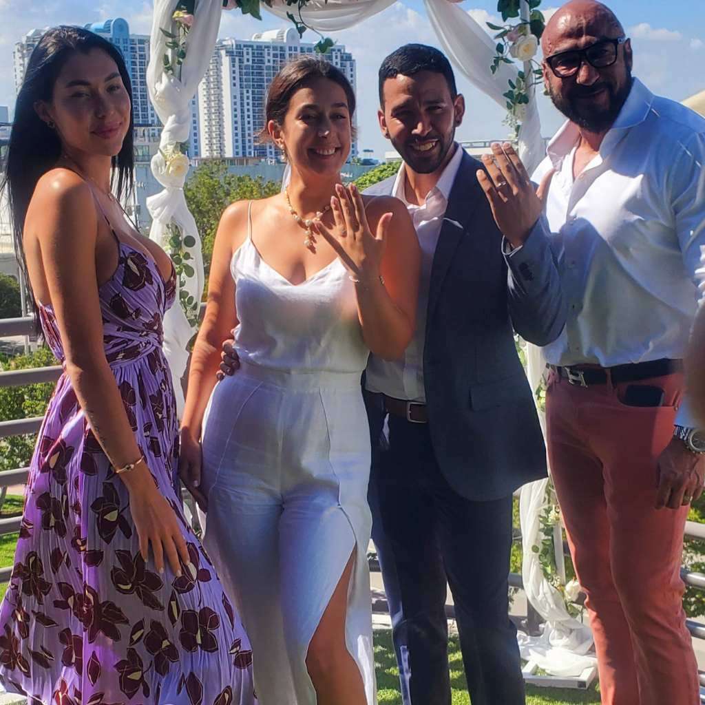 Four people posing outdoors in front of a decorated archway with city buildings in the background. Two of the people, a man and a woman in the center, are holding up their hands to display engagement rings. Everyone is smiling and dressed in summer attire, ready for a ceremony led by their marriage officiant.