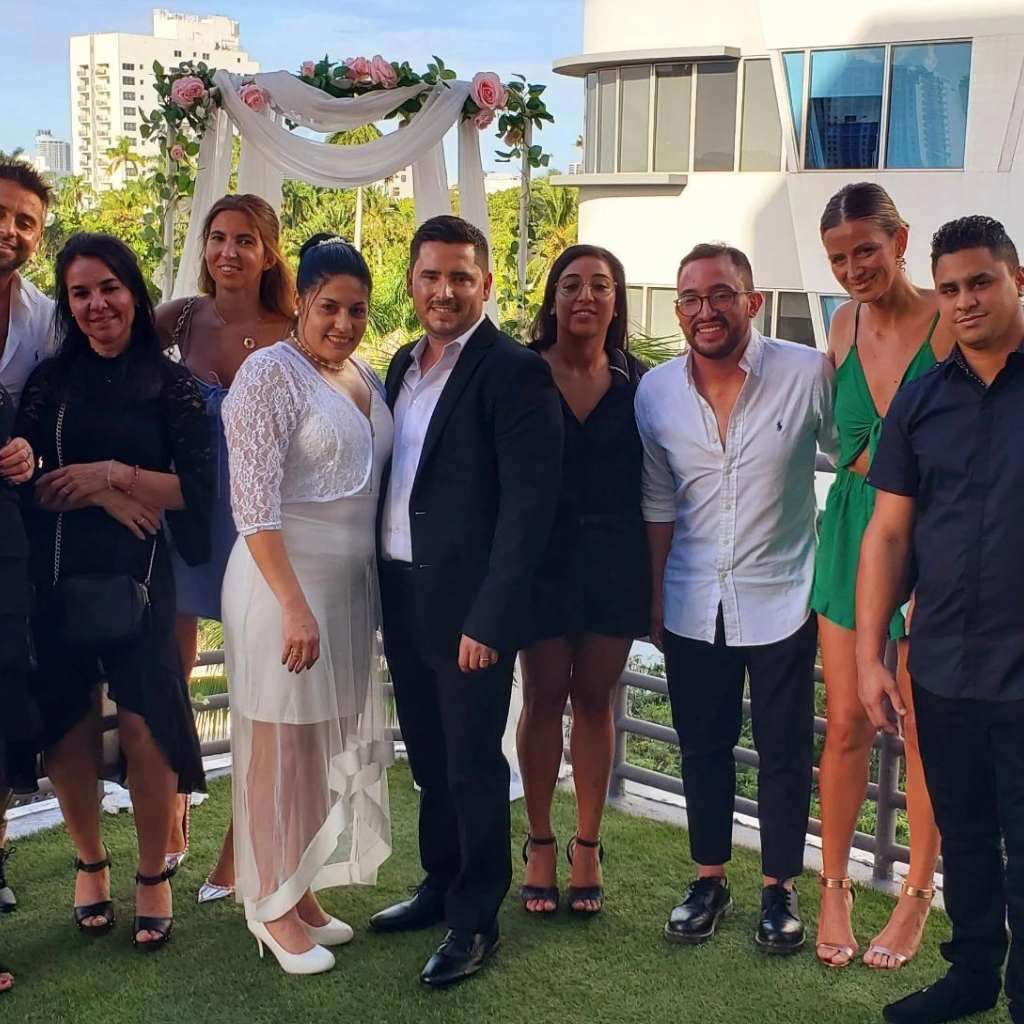 A group of people dressed formally and semi-formally are posing outdoors for a photo. In the center, a couple stands in front of a floral wedding arch. The background features modern buildings and lush greenery, capturing the essence of elegant family weddings.