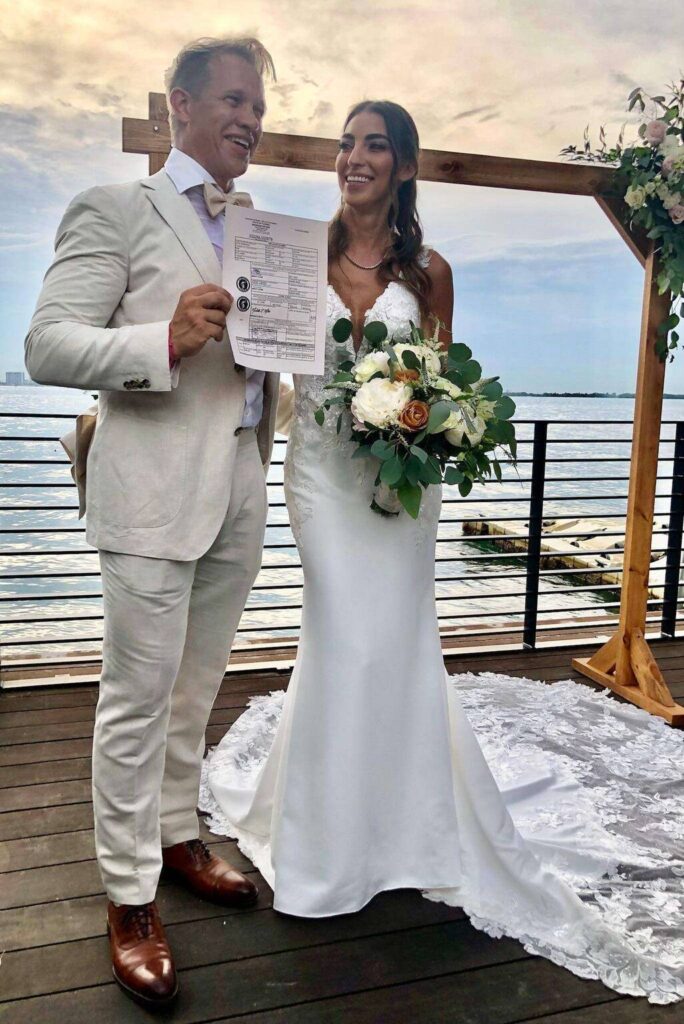 A smiling couple stands on a wooden deck by the water under a floral wedding arch, joined by an officiant for a wedding. The man holds up a document, while the woman in white holds a bouquet of white and peach flowers—celebrating their special day.