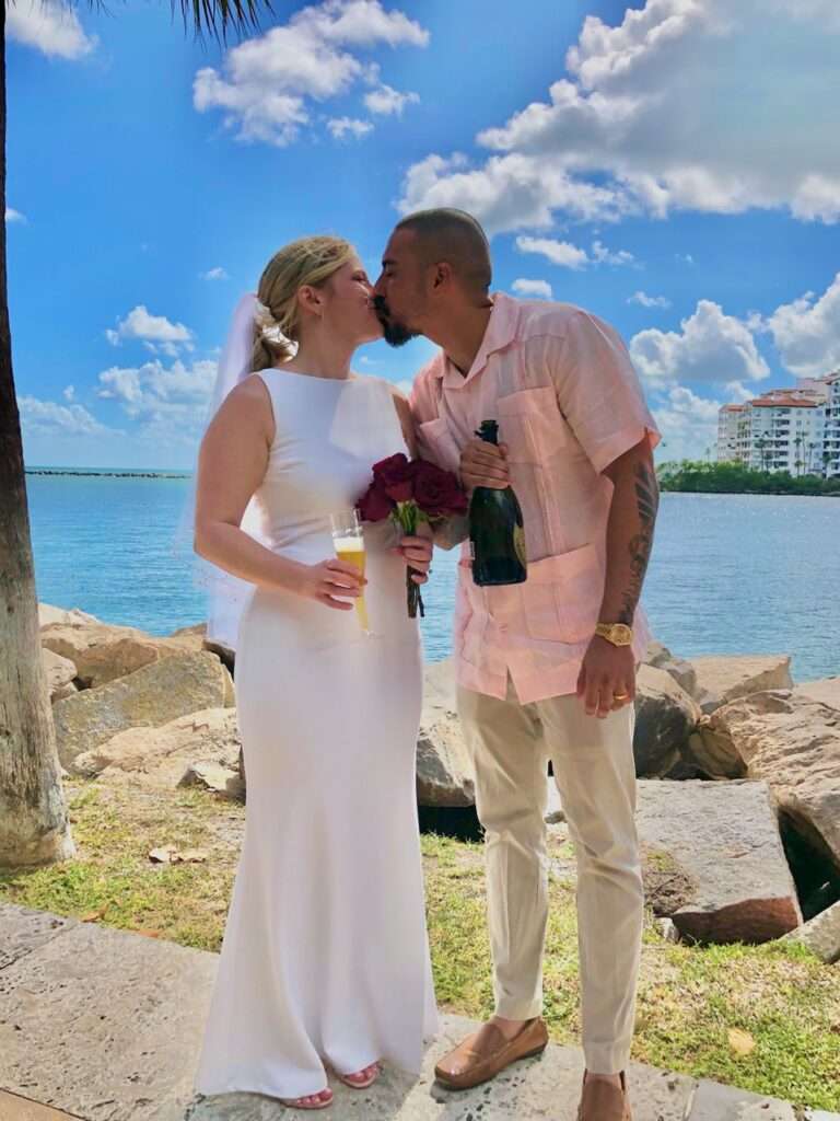 A couple sharing a kiss by the water on a sunny day. The woman, in a white dress and veil, holds a bouquet of red roses and a glass of champagne, while the man, in a light pink shirt and beige pants, holds a champagne bottle. Buildings and clouds dot the blue sky behind them. Thanks to mobile wedding services, their love story unfolds beautifully.