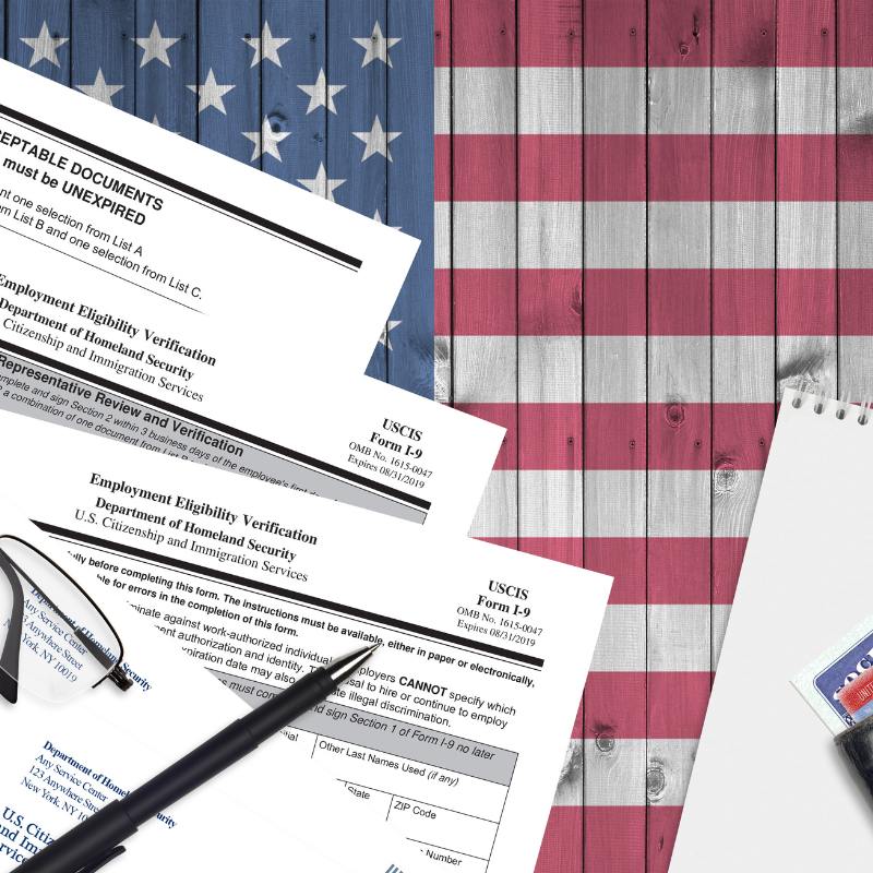 A desk displays several USCIS Employment Eligibility Verification Form I-9 papers, a pen, eyeglasses, a notepad, and a partially visible U.S. passport card. The background features a wooden American flag symbolizing employee authorization.