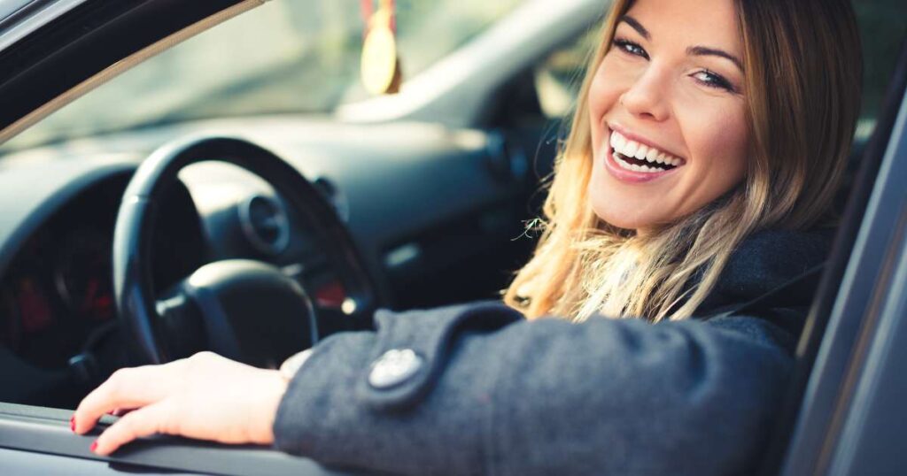 A woman with long blonde hair smiles broadly while sitting in the driver's seat of a car with the window rolled down. She is wearing a grey coat and looks cheerful and happy, perhaps on her way to provide mobile notary services from miami beach notary,. The interior of the car is modern with a dark-colored dashboard.