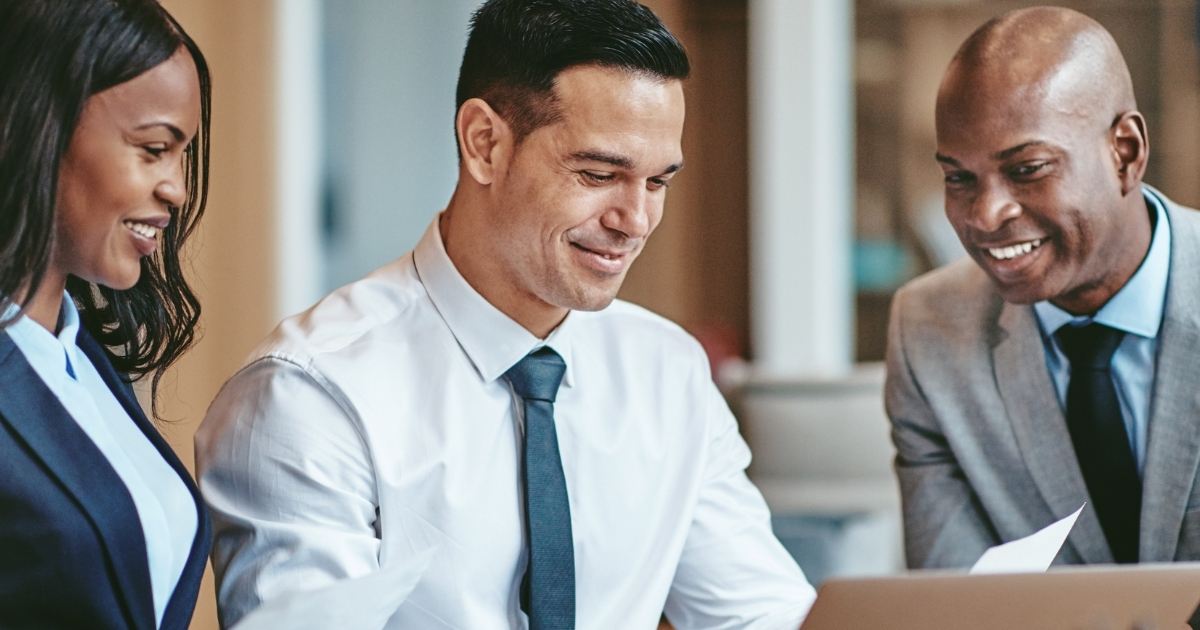 Three professionally-dressed people, including a legal assistant, are gathered around a laptop, smiling and engaged in conversation. Two men, one in a white shirt and tie, the other in a grey suit, and a woman in a navy blazer, are in a well-lit office environment.