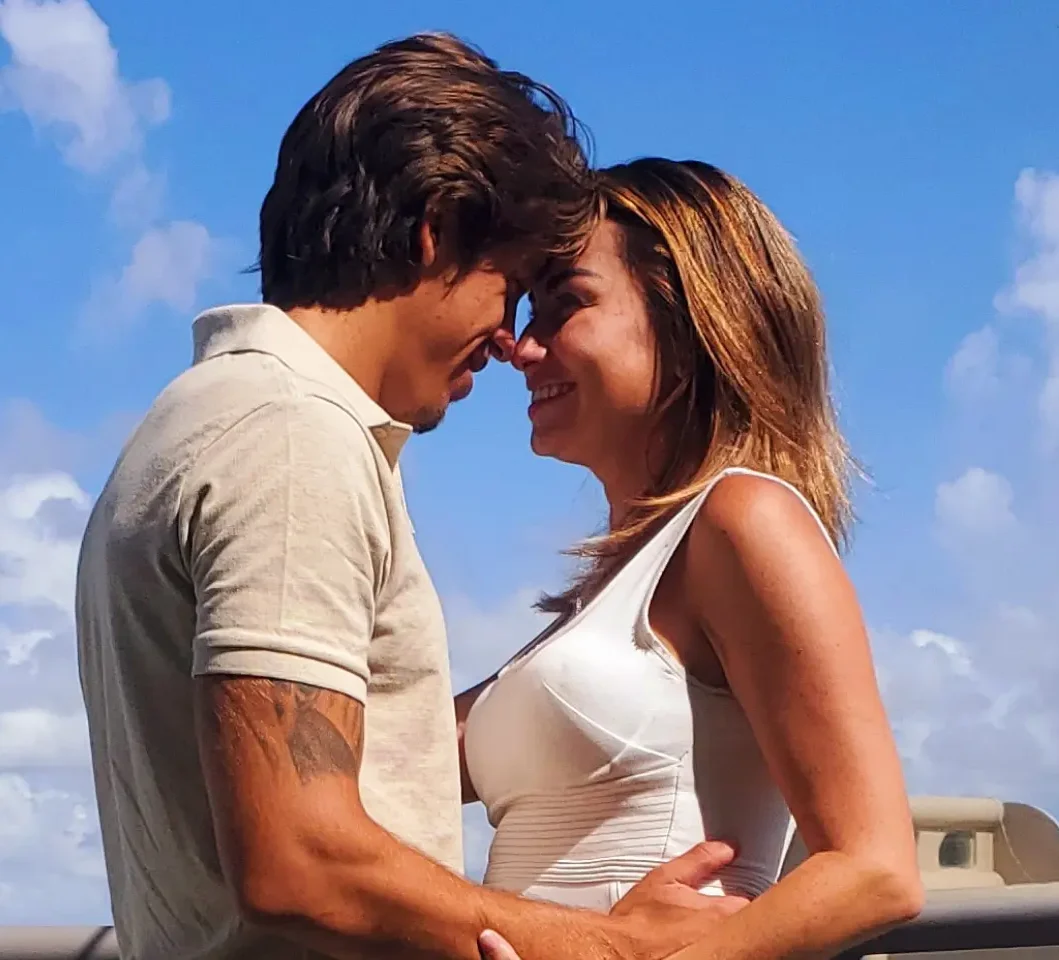 A couple stands close, smiling and gazing into each other's eyes against a backdrop of a bright blue sky with scattered clouds. The man, with dark hair and a tattoo on his left arm, wears a tan shirt, while the woman, with lighter hair, wears a white dress. A Miami beach notary marriage officiant stands nearby.