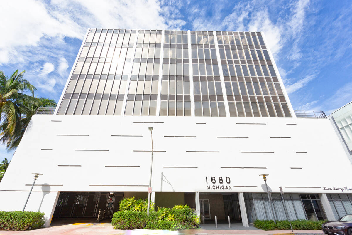 A modern building with a white facade and large vertical windows. The address "1680 Michigan" is displayed on the front. There are palm trees on the left side and a blue sky with scattered clouds in the background. A small black car is parked on the right.