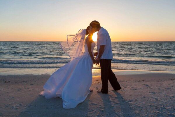 A bride and groom stand on a beach at sunset, holding hands and sharing a kiss. The bride wears a flowing white dress and veil, while the groom is dressed in a white shirt and dark pants. The sun sets over the ocean in the background, casting a warm glow as they exchange vows with using Miami Beach Notary Services