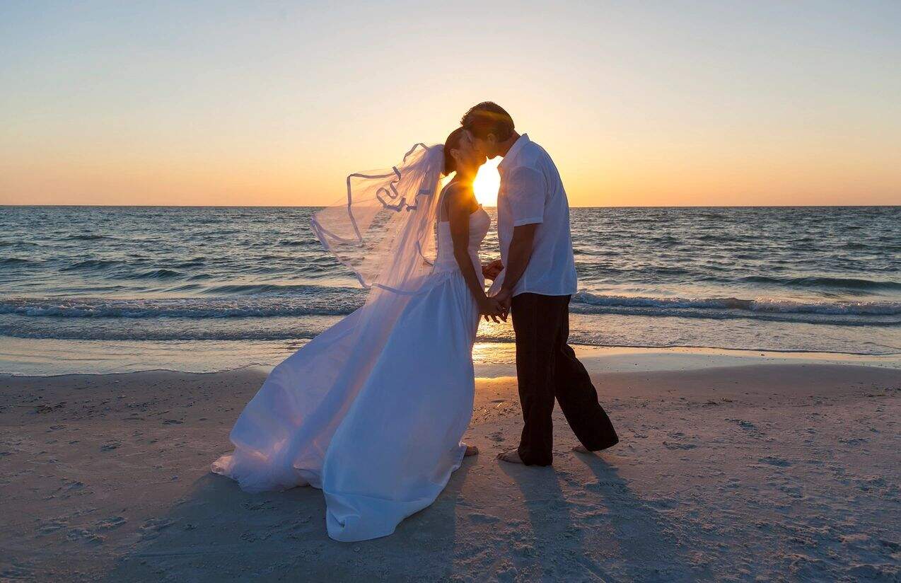 A bride and groom stand on a beach at sunset, holding hands and sharing a kiss. The bride wears a flowing white dress and veil, while the groom is dressed in a white shirt and dark pants. The sun sets over the ocean in the background, casting a warm glow as they exchange vows with using Miami Beach Notary Services