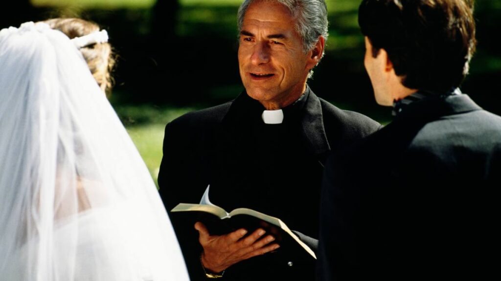 A bride and groom stand face to face outdoors, with a wedding officiant positioned between them. The officiant holds an open book, presumably a Bible, and appears to be speaking. The bride wears a veil, and the groom is in a black suit. Trees and greenery are visible in the background.