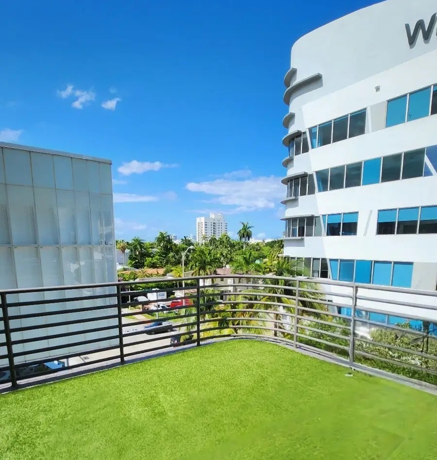 View from a balcony with artificial grass flooring and a modern metal railing. The scene features a cityscape with a mix of white and glass buildings, palm trees, parked cars, and a bright blue sky with scattered clouds. Wedding Officiant