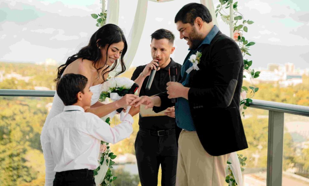 A bride and groom stand on a balcony exchanging rings during a wedding ceremony. A young boy hands the ring to the bride as the marriage officiant in black holds a microphone between them. The Wynwood balcony is adorned with green vines, and trees are visible in the background under a cloudy sky.