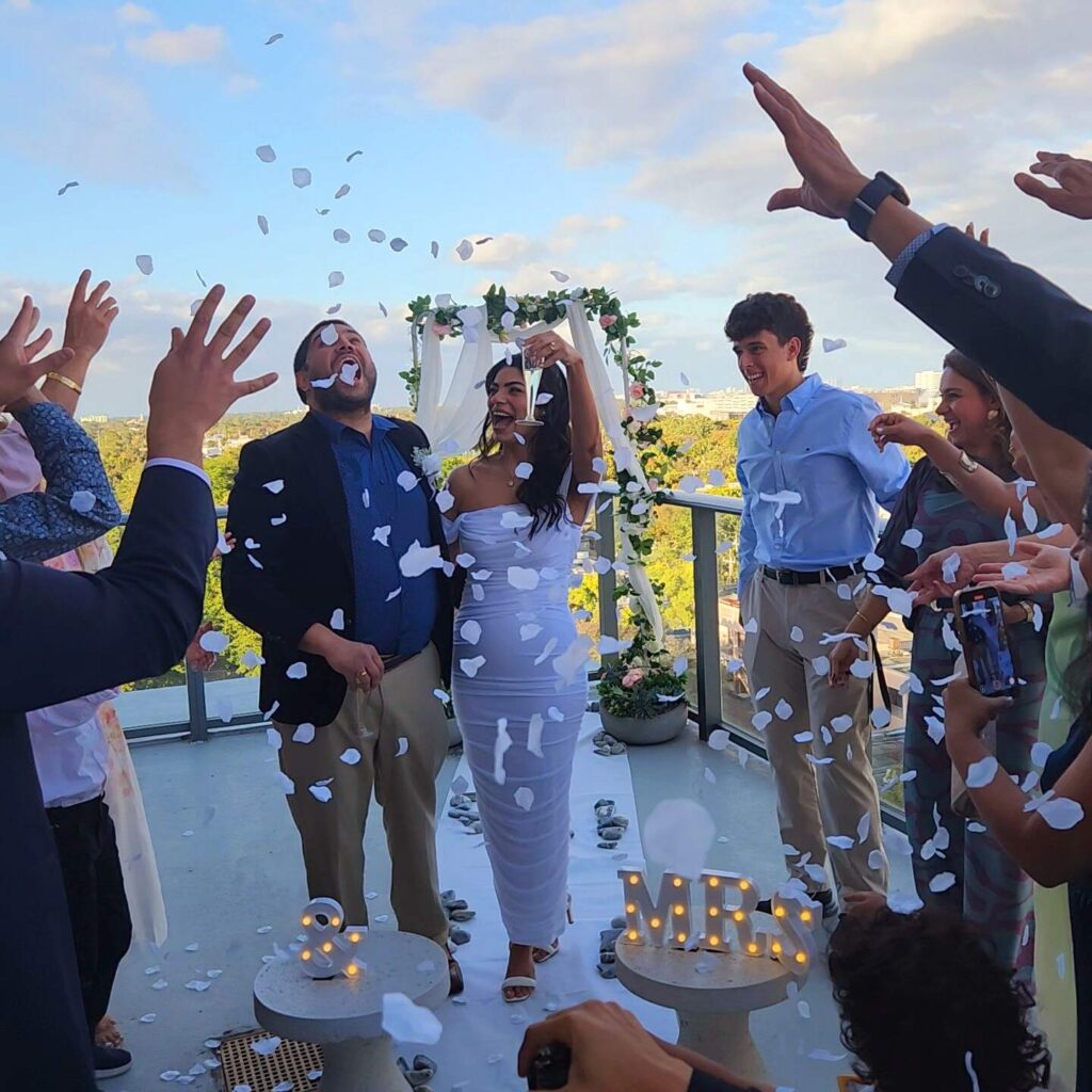 A couple, smiling under a floral arch, surrounded by well-dressed guests on a balcony. With Miami Beach notary services ensuring every detail is perfect, the bride in white holds her bouquet while guests throw petals. Light-up "MR." and "&" signs add to the romantic ambiance.