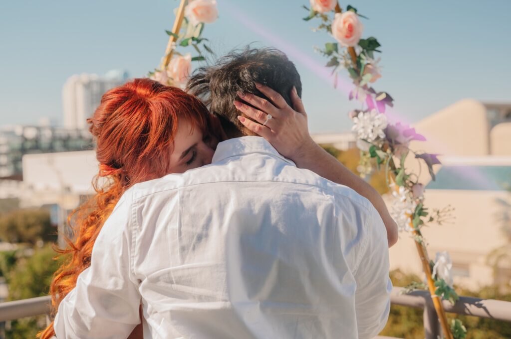 A woman with long red hair hugs a man outdoors under a floral arch. She holds his head gently as they embrace. Both wear white shirts. Pink flowers and greenery decorate the arch, and buildings are visible in the blurred background on a sunny day.