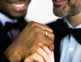 A close-up of two people holding hands, both wearing suits and bow ties. One person has darker skin and the other has lighter skin with a beard. The person with darker skin is wearing a gold ring. They are both smiling, celebrating their same-sex marriage.