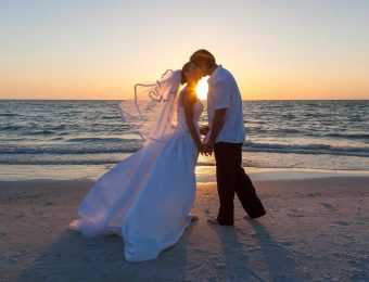 A bride and groom stand on a beach at sunset, holding hands and sharing a kiss. The bride wears a flowing white dress and veil, while the groom is dressed in a white shirt and dark pants. The sun sets over the ocean in the background, casting a warm glow as they exchange vows with using Miami Beach Notary Services